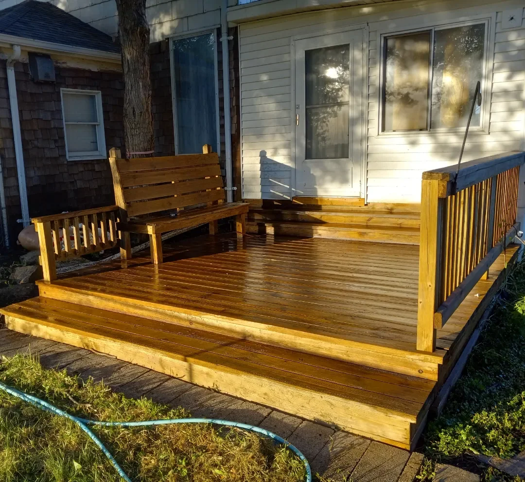 Power washed wooden deck with a seating bench, removed dirt and mold from wood, attached to a white house with a door and window, during late afternoon sunlight.
