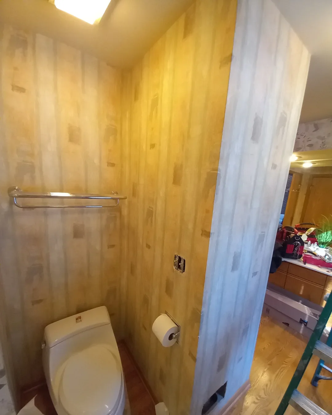 A small bathroom with unfinished wooden walls, a toilet, a metal towel bar, and a toilet paper holder, with a doorway revealing a cluttered room beyond.