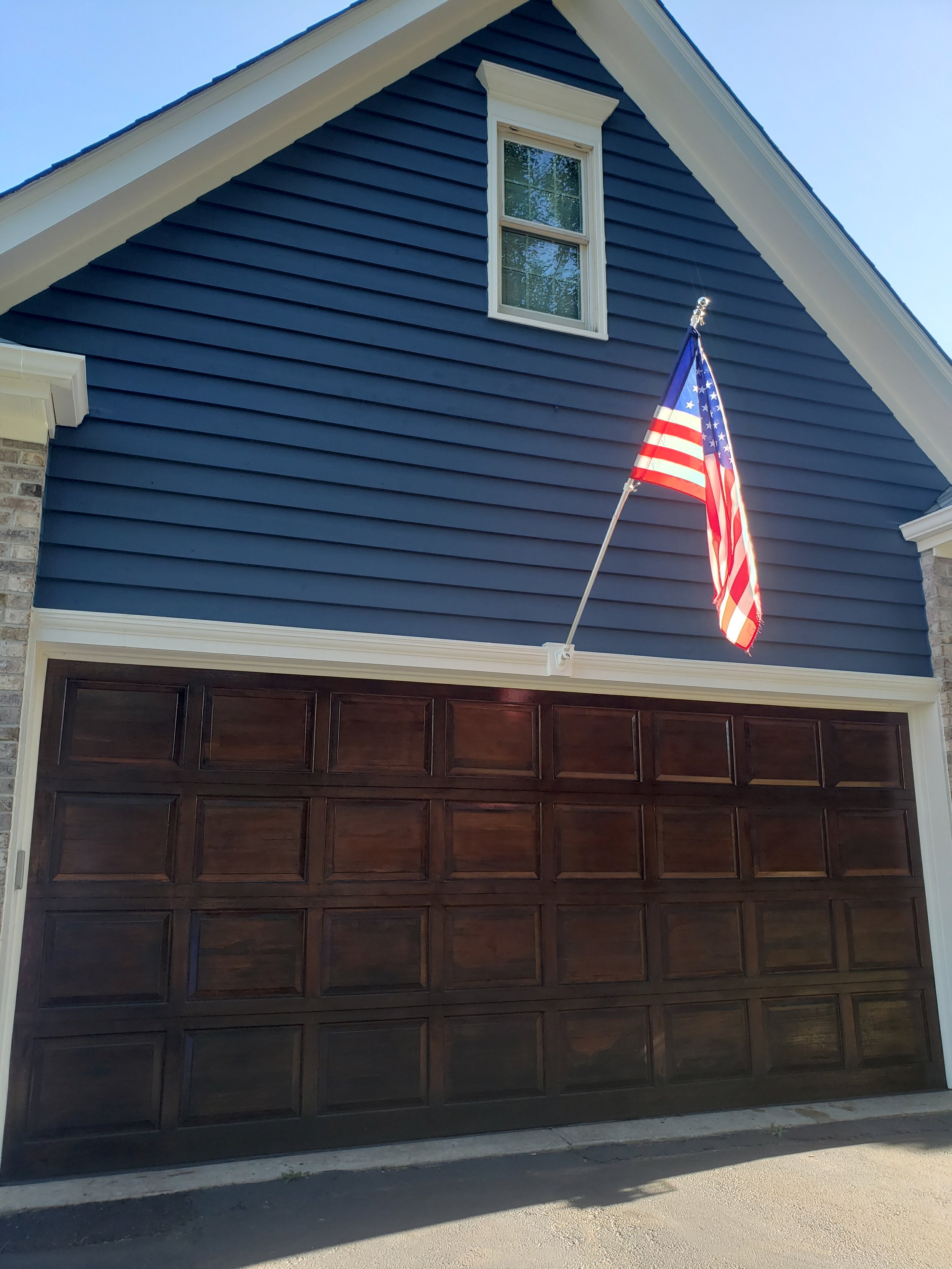 Restored dark brown wooden garage door, with painted blue siding on the upper part, a small window in the peak of the roof, and an American flag mounted on the blue siding, blowing in the breeze.