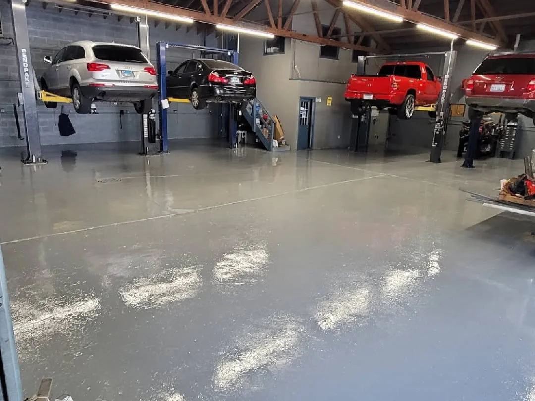 An indoor car repair shop with four cars on hydraulic lifts, including a silver SUV, a black sedan, a red pickup truck, and a red SUV. The floor appears wet or freshly coated, reflecting the overhead lighting.