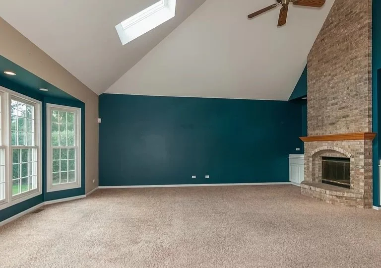 Living room with blue accent wall, large windows, carpet flooring, a brick fireplace, a ceiling fan, and a skylight.