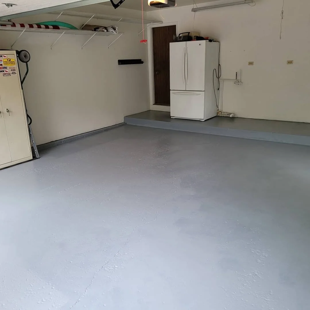 Garage with epoxy sealed flooring a white refrigerator, a wall-mounted shelf, and a storage cabinet, with concrete flooring and a slightly raised area near the door.