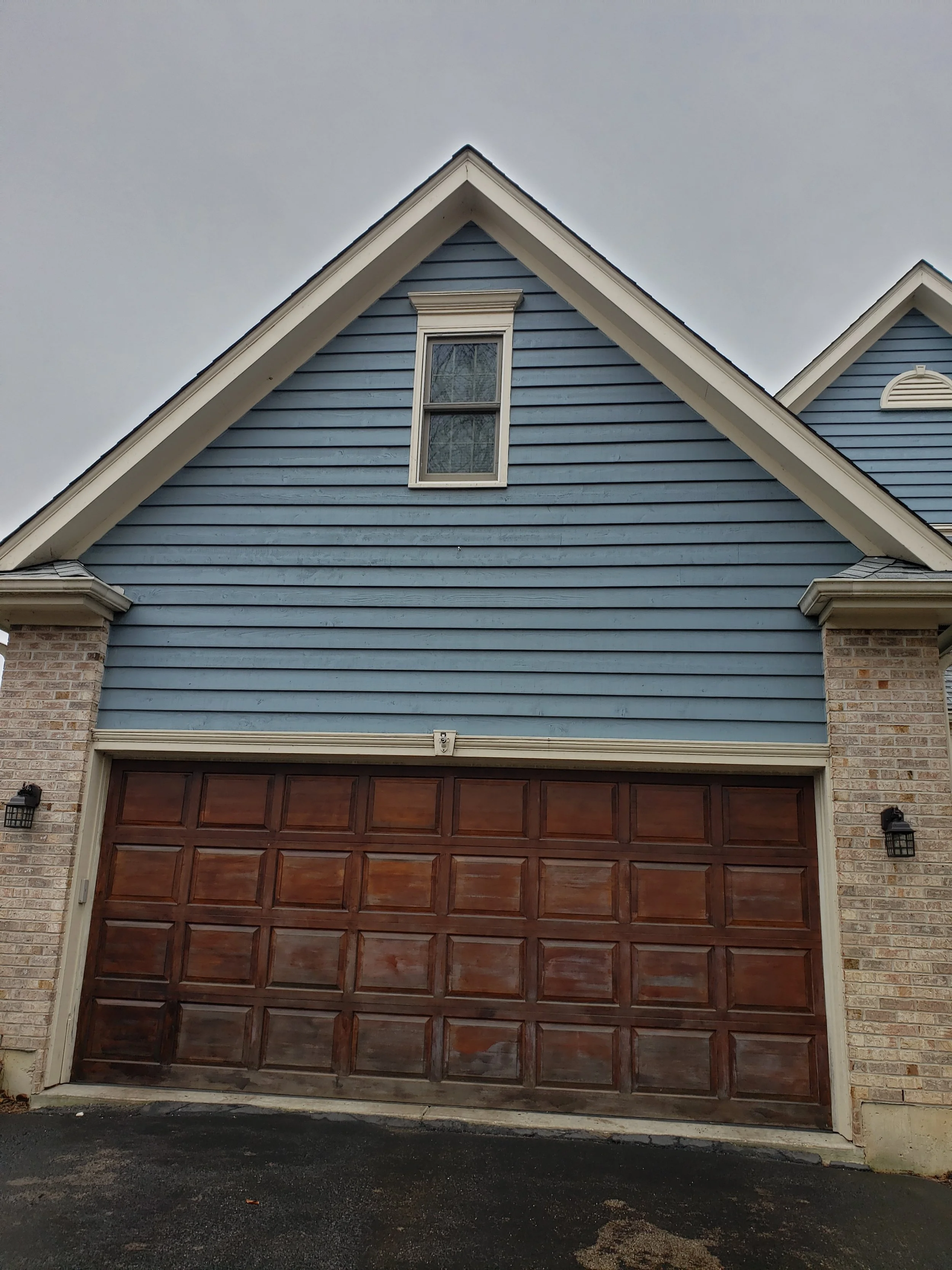 Front view of a house with a weathered wooden garage door, and faded blue wood siding, brick columns, and a single window on the upper level under a gray sky.