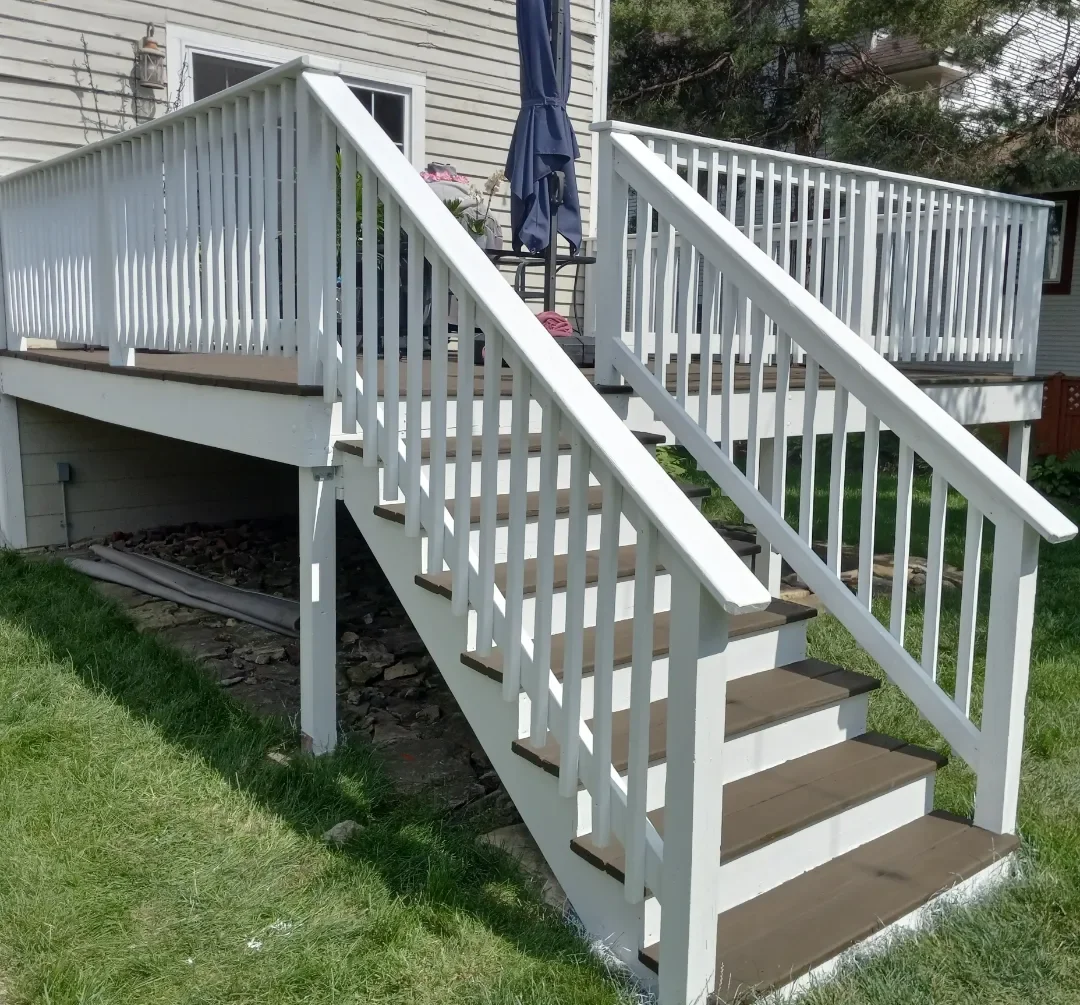 A wooden deck repainted white to brighten up the living space with stairs and railings attached to a house, with a small patio area and an umbrella on top of the deck.