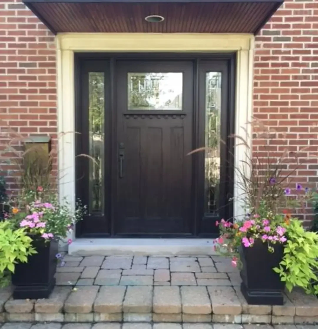Dark brown front door entrance with sidelights and a transom window, flanked by potted flowers on a brick walkway.