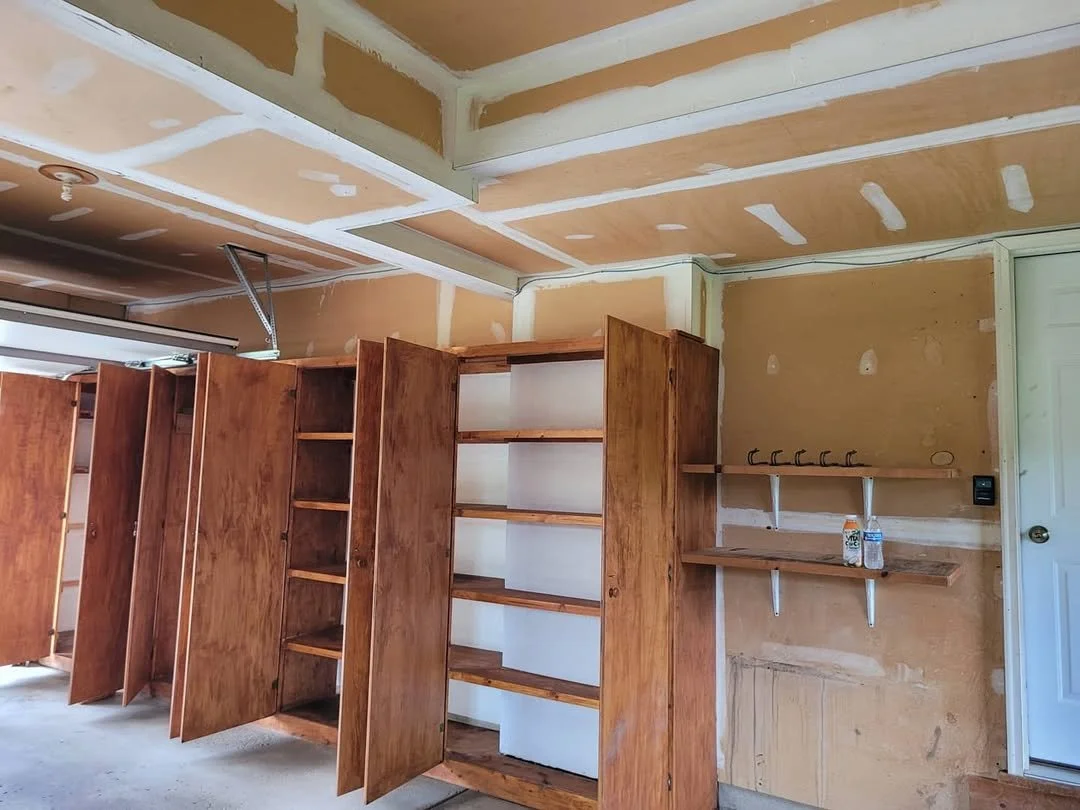 Unfinished garage walls with wooden shelves, cabinets, ceiling.