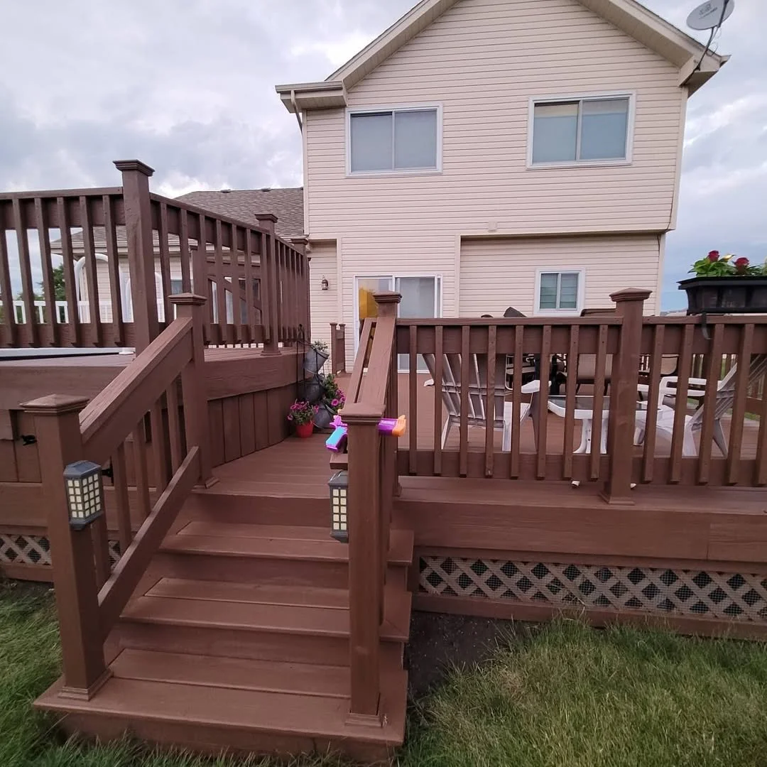 A repainted in dark brown wooden backyard deck with stairs, surrounded by a wooden fence, with outdoor furniture and potted flowers, attached to a two-story beige house with multiple windows.