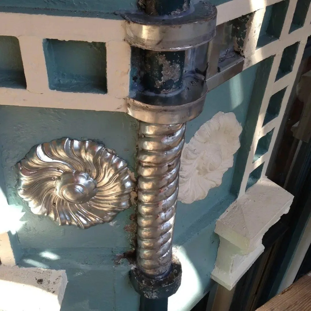 Close-up of a restored and added decorative metal spiral pipe connected to a structure with ornate metallic details and some brickwork, part of a construction or renovation site.
