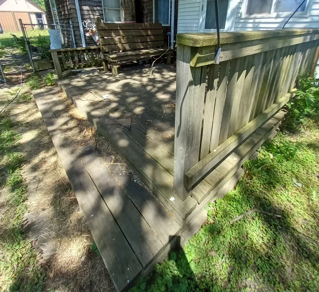 Weathered and dirty wooden deck with a seating bench,  and a small wooden railing, part of a house's backyard, surrounded by grass and small plants.