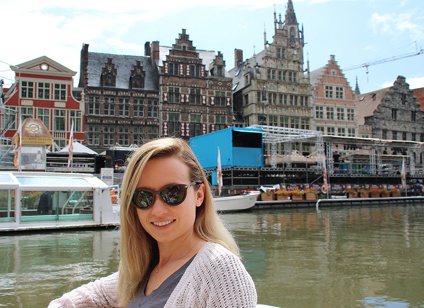Smiling woman with sunglasses taking a boat ride along a canal in front of historic European-style buildings.