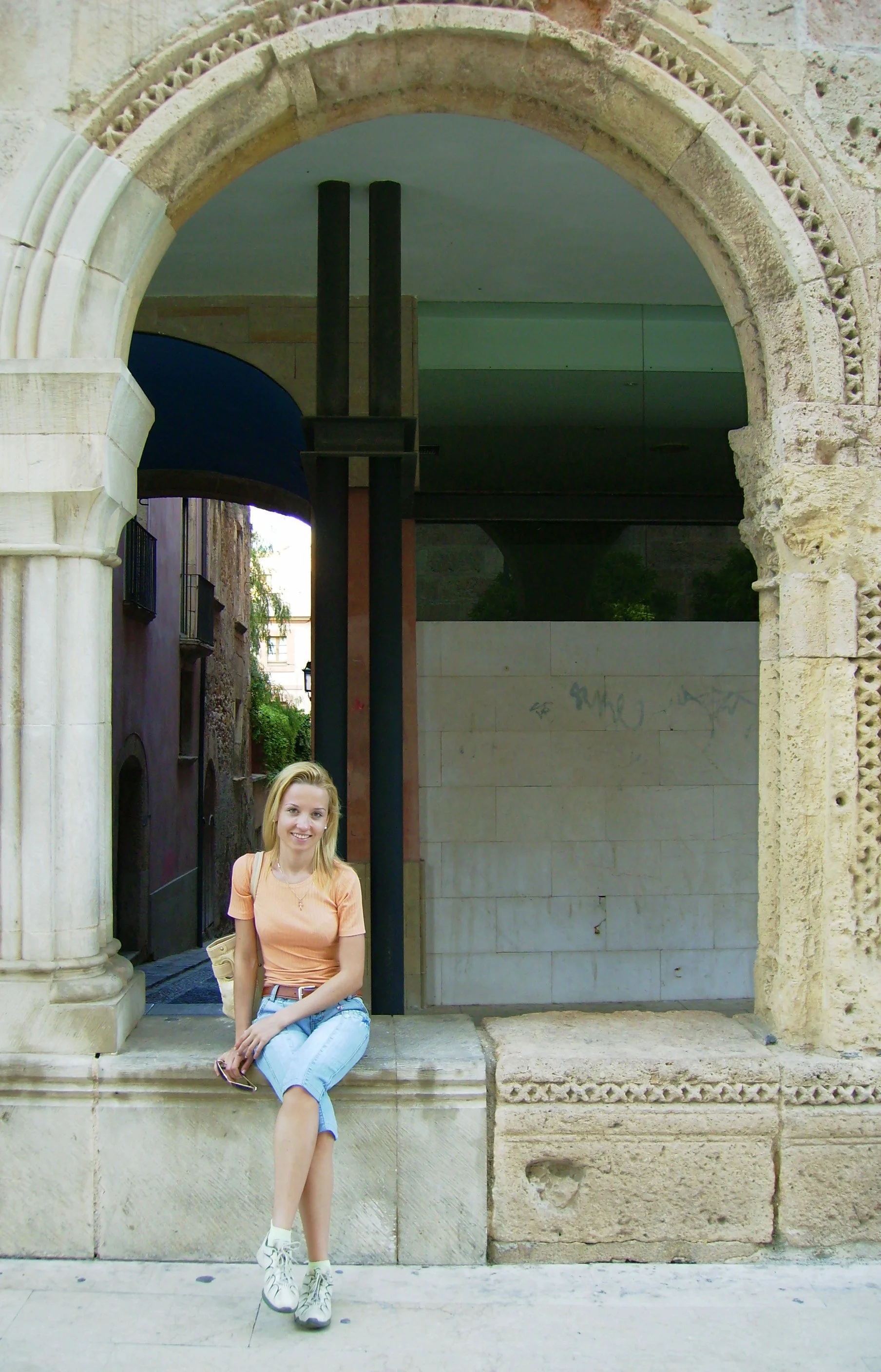 Young woman with blonde hair, wearing a peach-colored t-shirt, light blue jeans, and white sneakers, sitting on a stone step in front of an ornate stone archway in an urban setting.