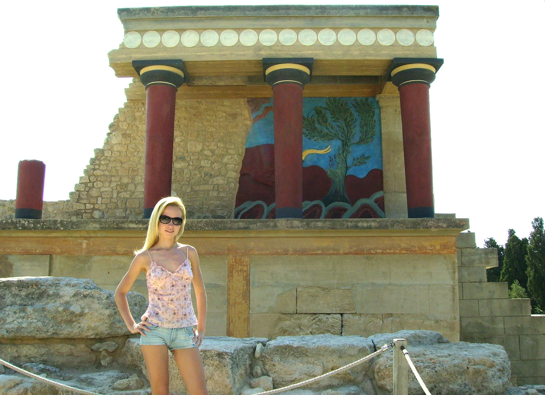 A young woman standing outdoors in front of ancient ruins with a colorful mural of a tree and mountains painted on a wall behind her. The woman is wearing sunglasses, a floral tank top, and denim shorts.