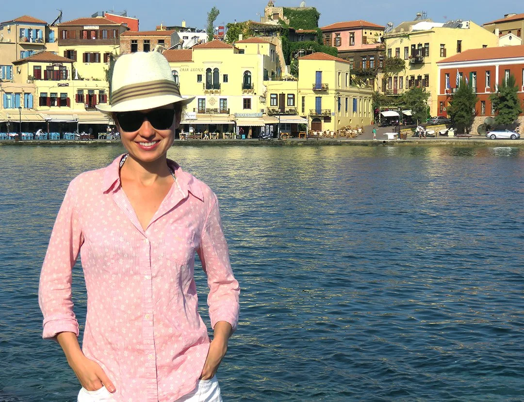 A woman smiling, wearing sunglasses and a white fedora hat, standing in front of a body of water with colorful buildings on a hillside in the background.