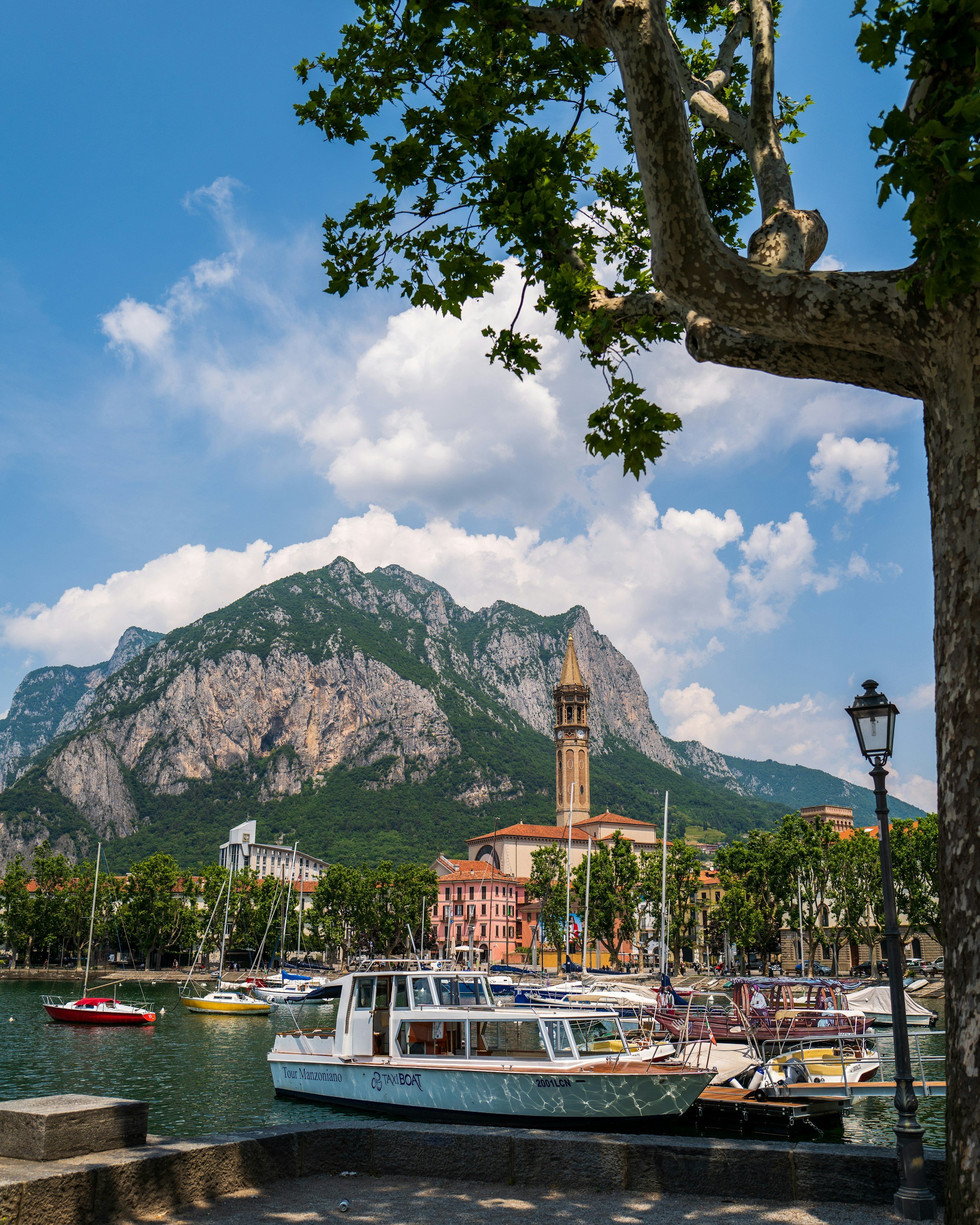 View of boats docked at a marina in a town with colorful buildings, a church tower, and green trees in front of mountainous scenery under a partly cloudy sky.