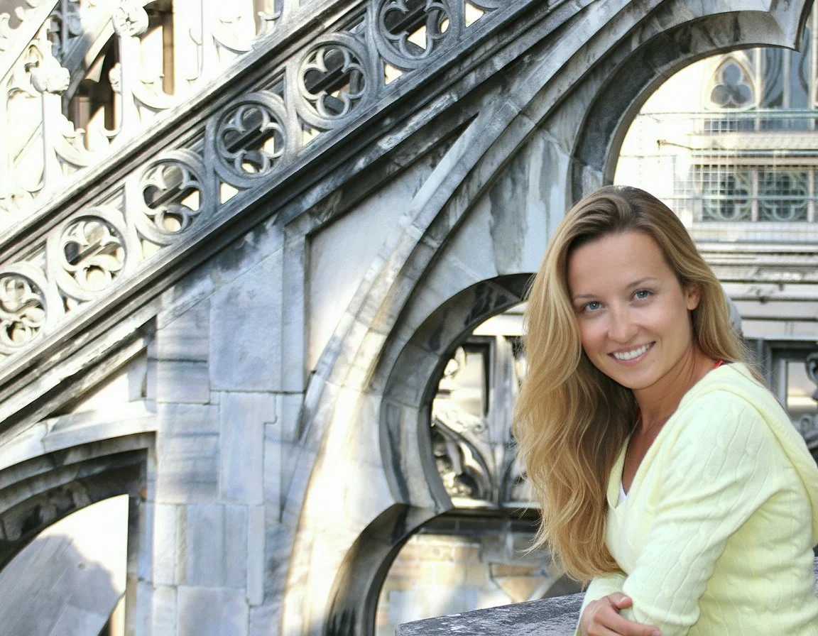 A smiling woman with long, blonde hair wearing a light yellow top, sitting on the steps of a stone and metal ornate bridge in an urban setting.