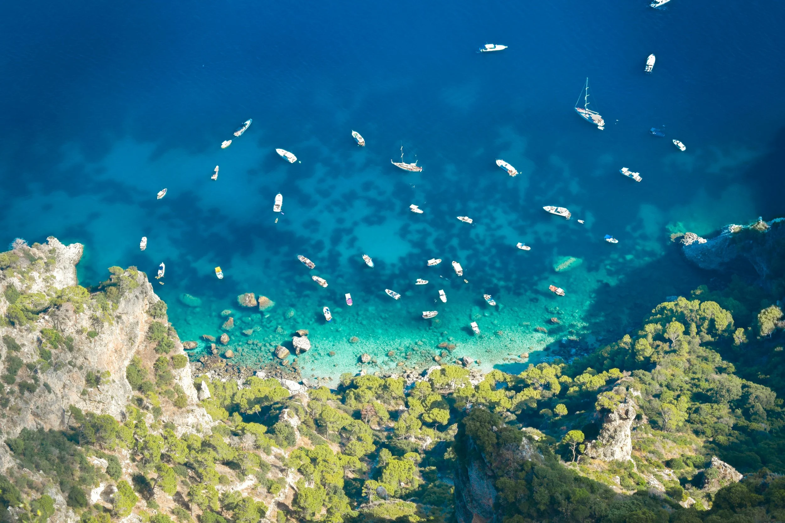 Aerial view of a turquoise bay with numerous boats anchored near a rocky, forested coastline.