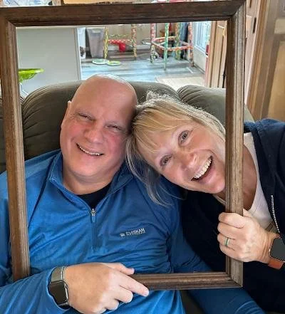 Smiling couple sharing a joyful moment together while holding a wooden picture frame around their faces.
