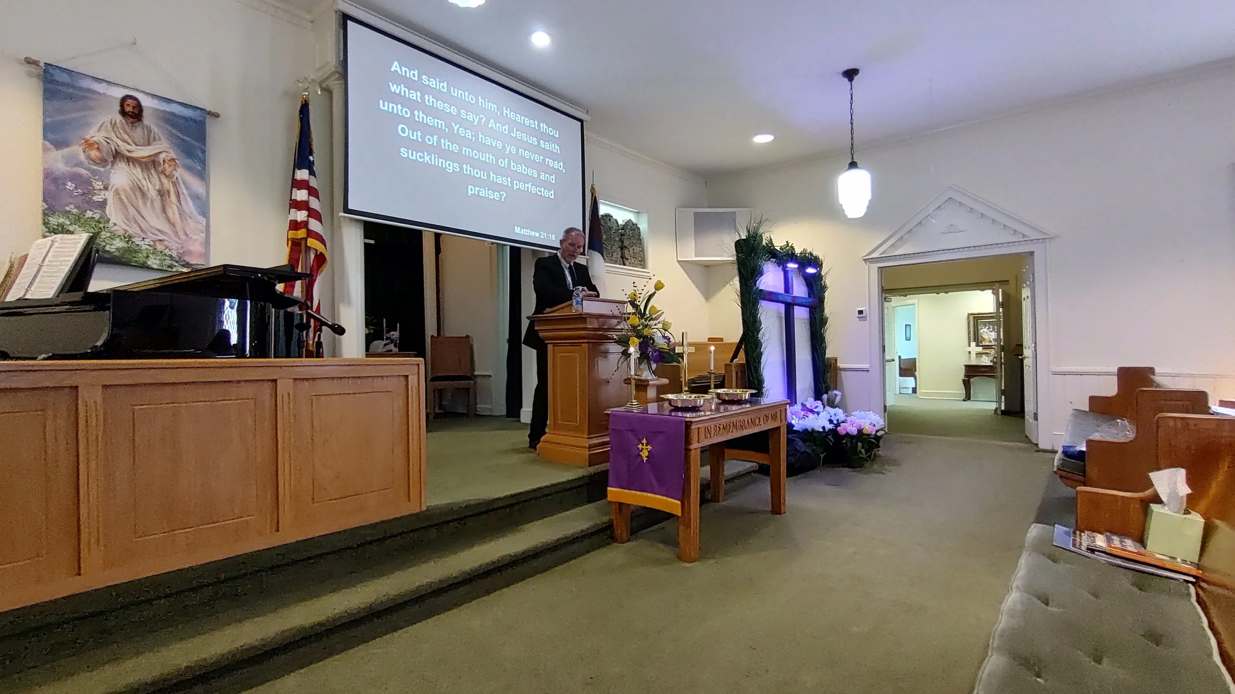 Inside a church with wooden pews and a stage. A man stands at a pulpit, and a large screen displays a Bible verse. There are American and church flags, floral arrangements, and religious decor.