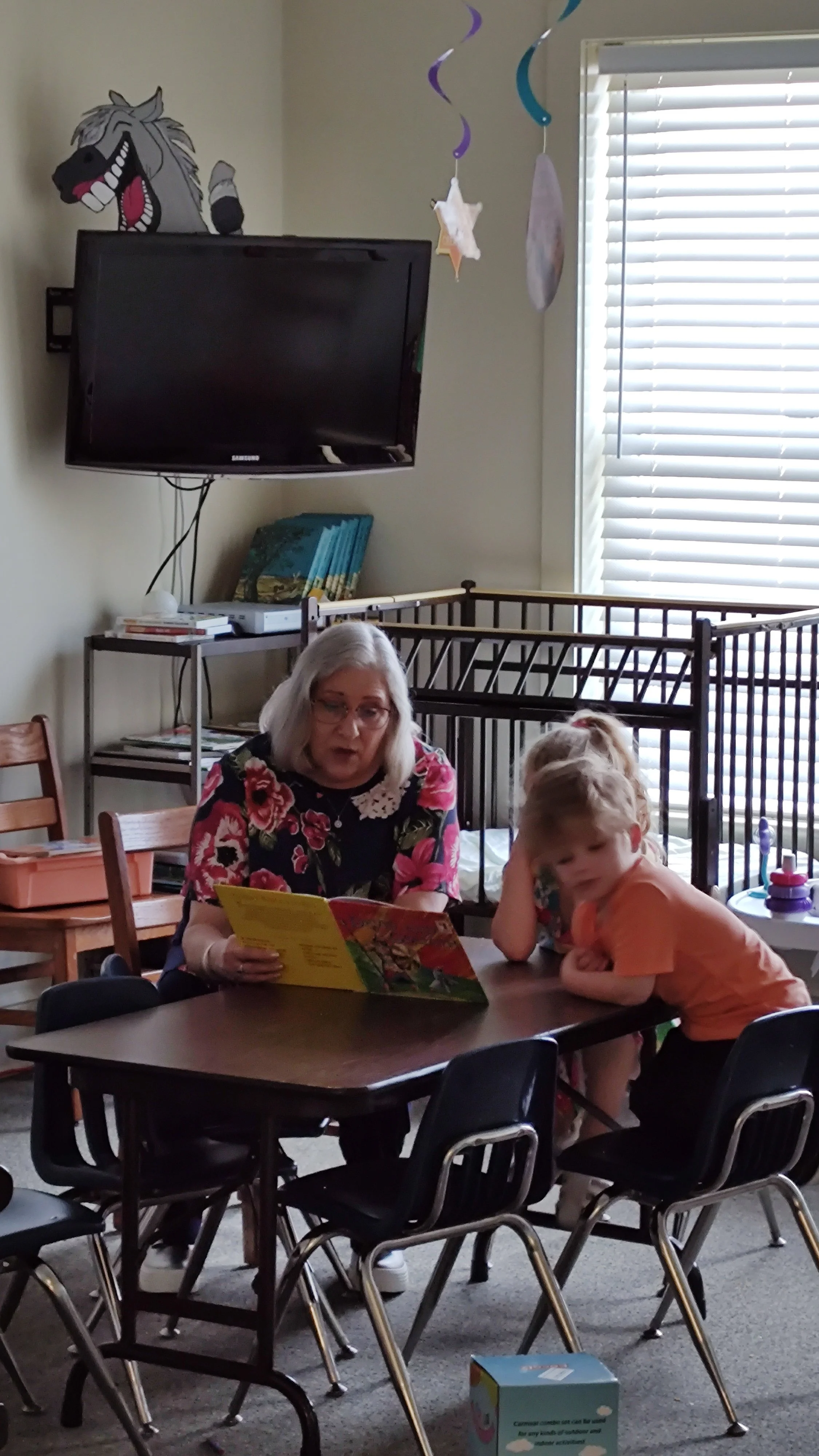 An elderly woman reading a colorful book to two young children at a table in a room decorated with hanging paper shapes, a mounted TV, and a metal crib in the background.