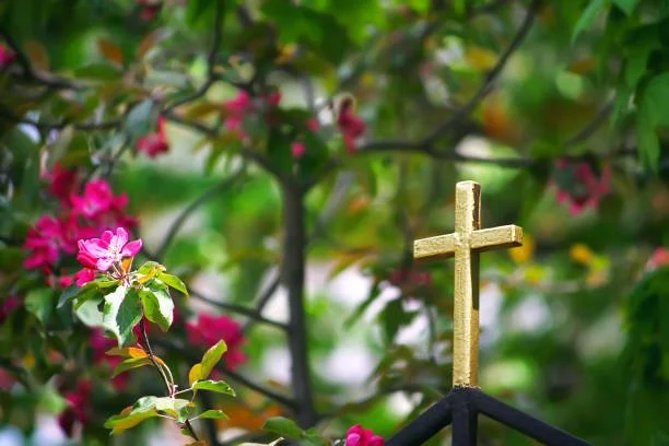 A gold Christian cross on a black fence with pink flowers and green foliage in the background.
