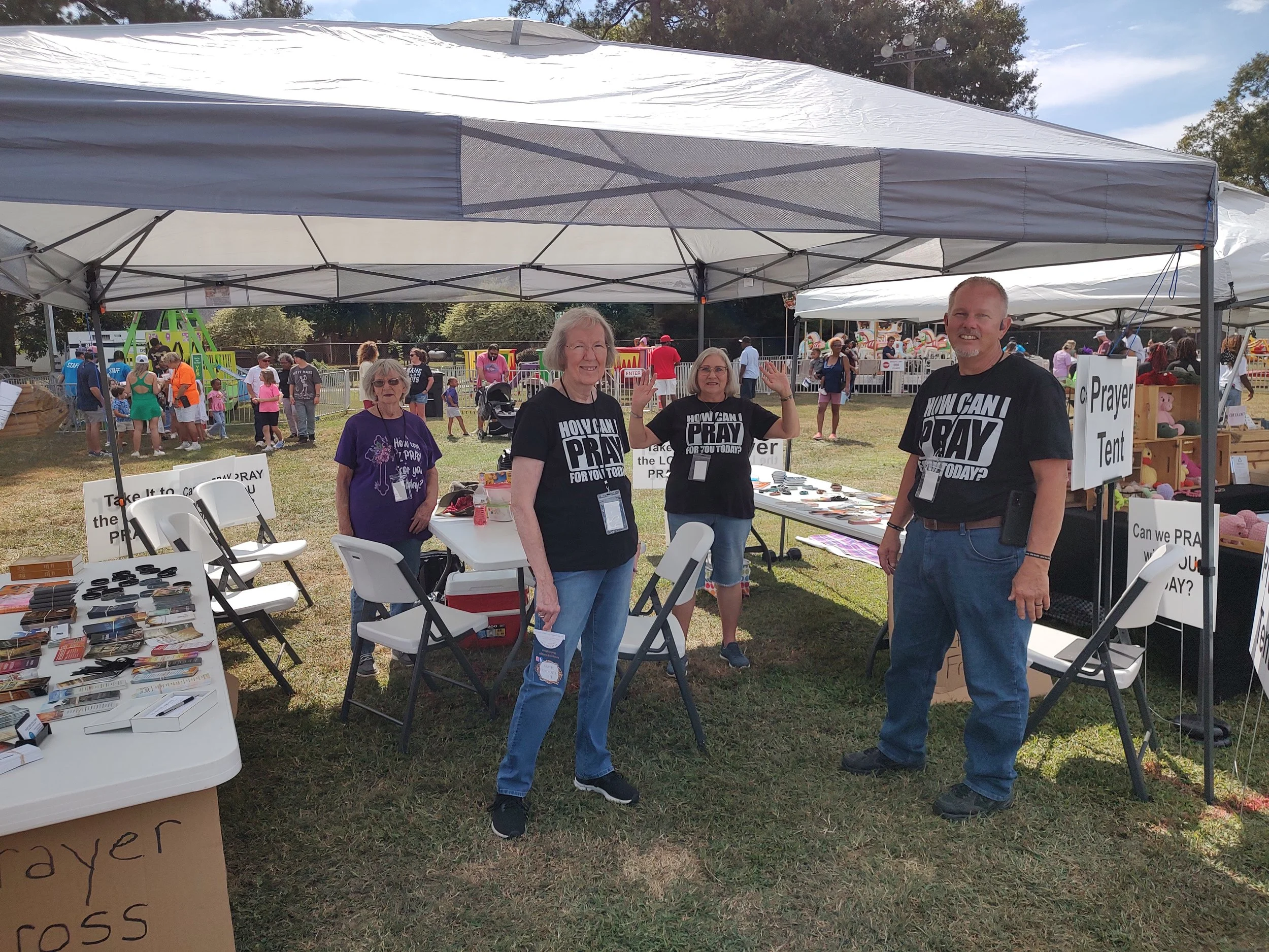 People standing under a white canopy at a prayer tent booth at an outdoor event with children’s rides in the background.