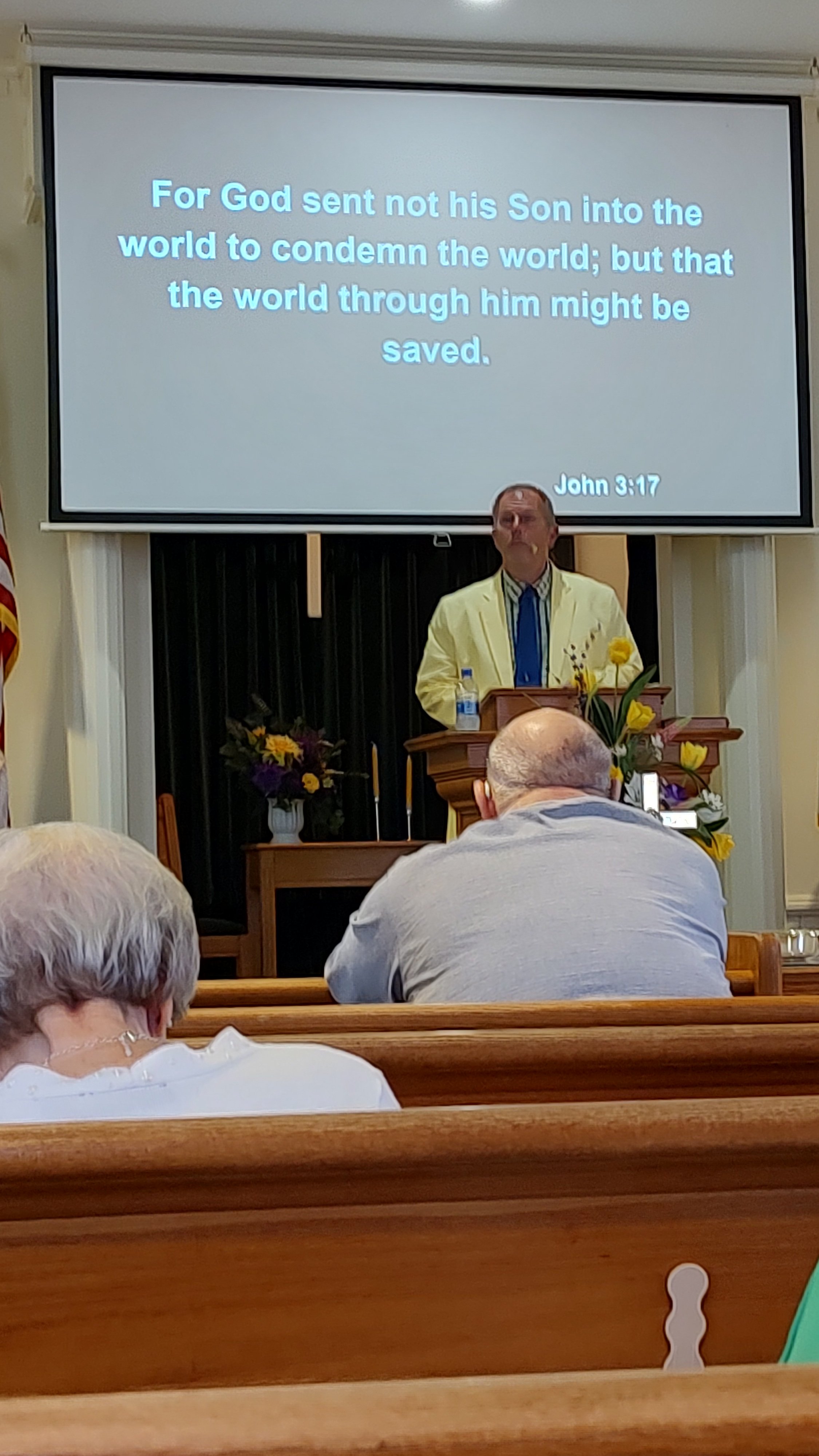 A man in a yellow blazer standing at a pulpit during a church service as a screen behind him displays a Bible verse from John 3:17 about Jesus being sent to save the world, with attendees seated in pews.