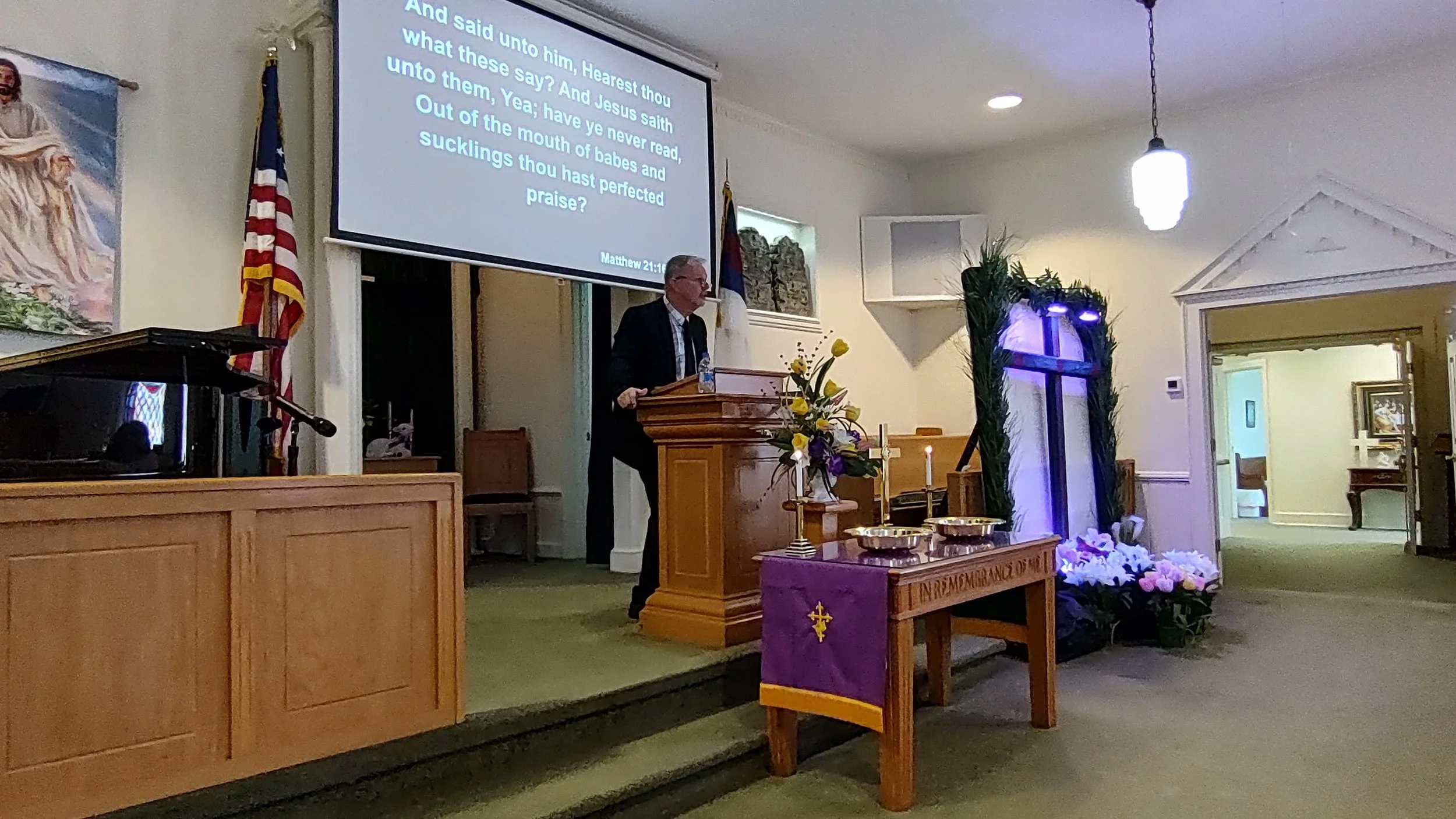 A church service with a pastor standing at a pulpit, holding a Bible. The background shows a large screen projecting a Bible verse from Matthew 21:16. The church is decorated with flowers, candles, and greenery, with American and Christian flags nearby. The interior has soft lighting and an open door leading to another room.