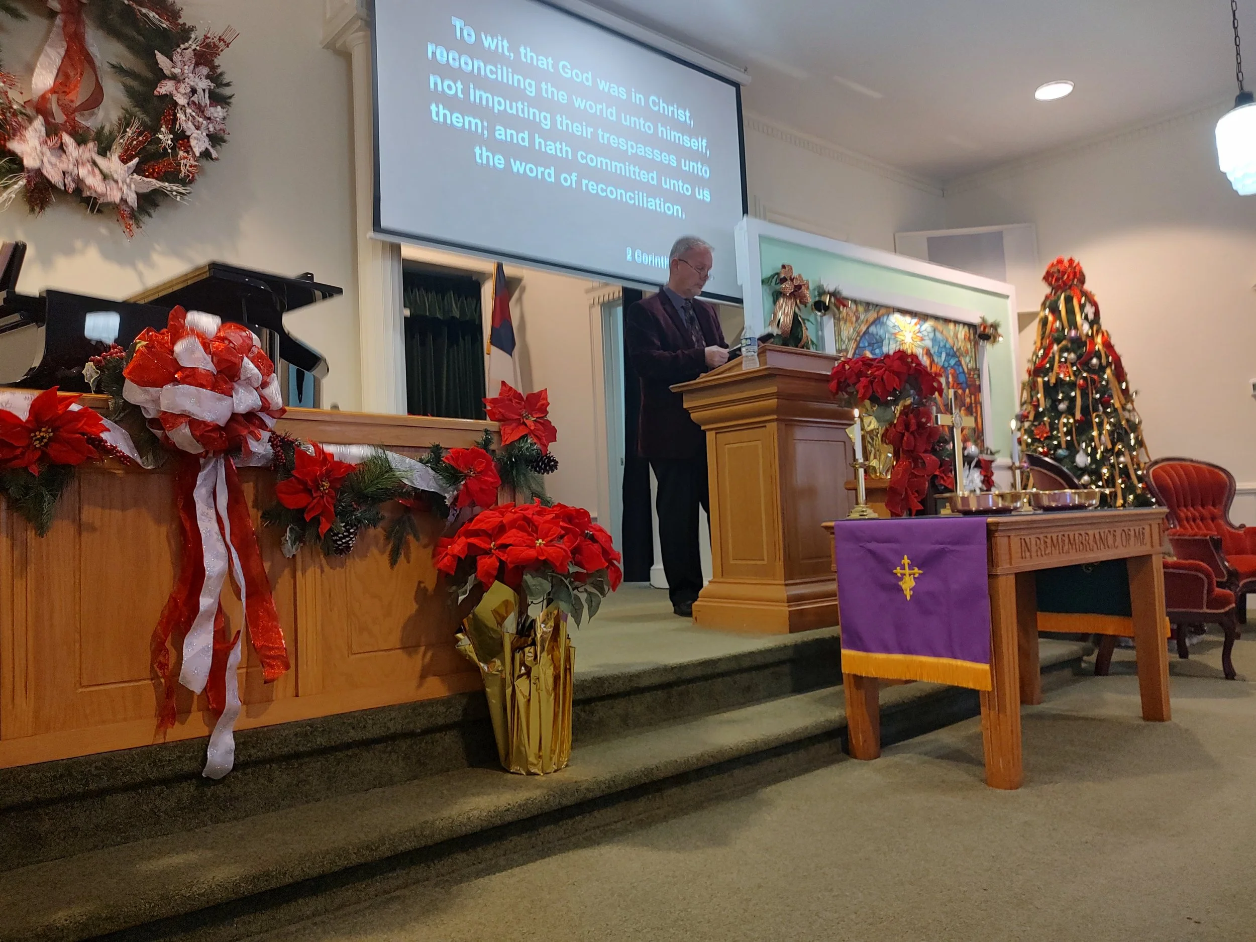 A church decorated with Christmas flowers, ribbons, and poinsettias, with a Christmas tree on the right, a man standing at a podium, and a large screen displaying a biblical passage behind him.