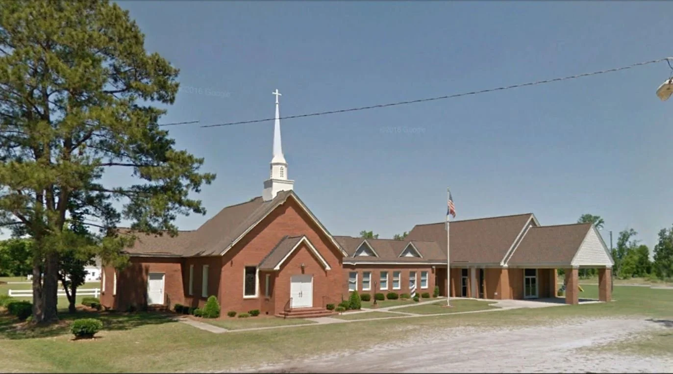 A church with red brick walls, a tall white steeple, and a flagpole in front. The building is surrounded by green grass and a few trees.