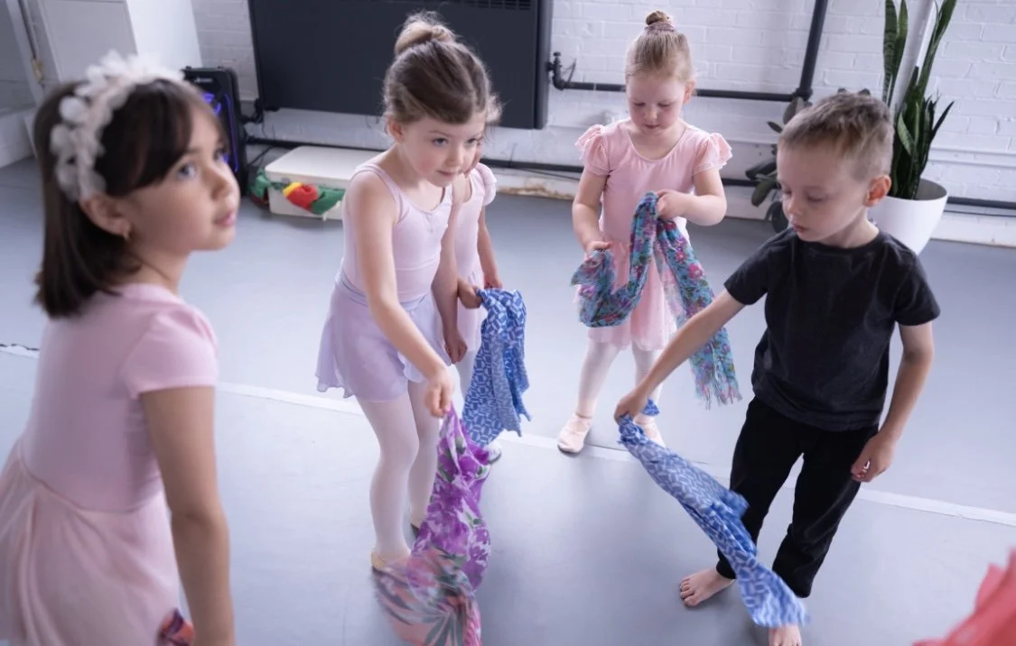 Four young children, three girls and one boy, are standing in a dance studio holding colorful scarves, possibly preparing for a dance or play activity.