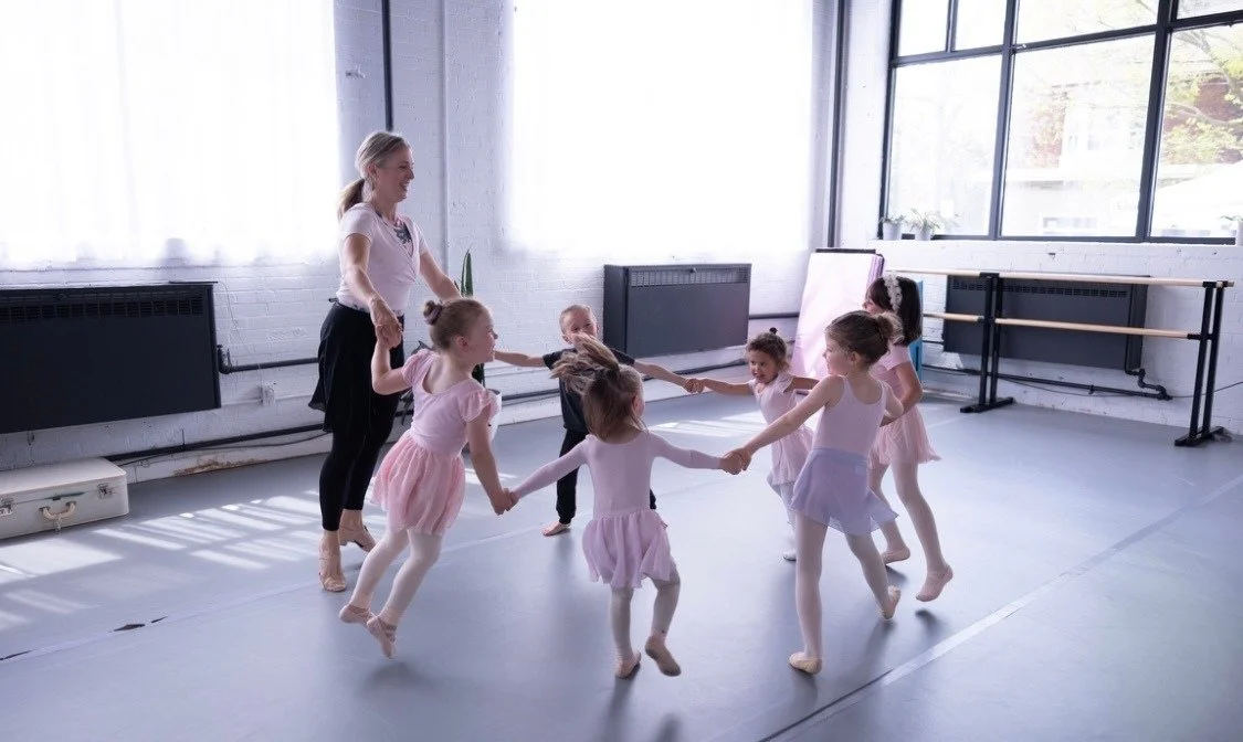 A dance class with young girls in ballet tutus holding hands in a circle, led by an instructor in a dance studio with large windows and ballet barres.
