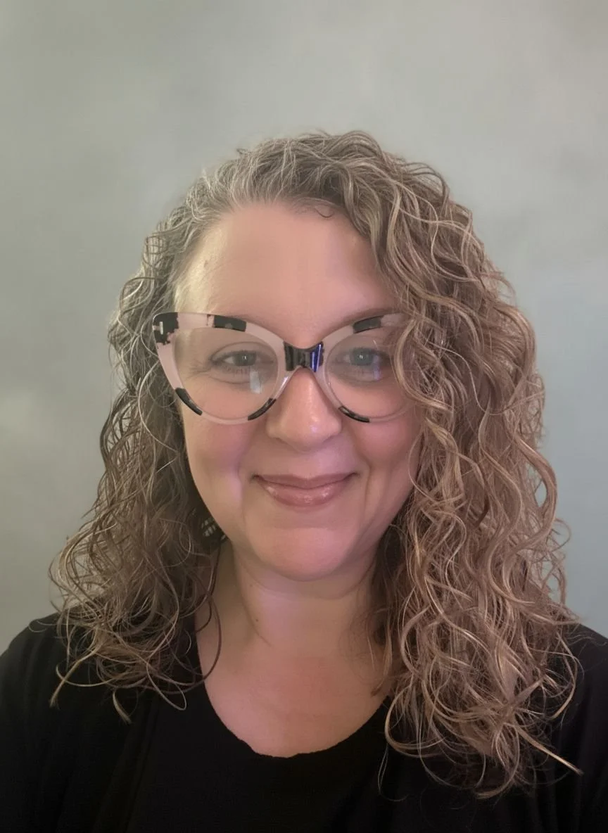 A woman with curly blonde hair wearing glasses and a black top, smiling at the camera against a plain background.