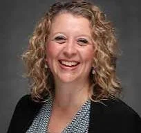 A woman with curly blonde hair smiling, wearing a black blazer and a patterned shirt.