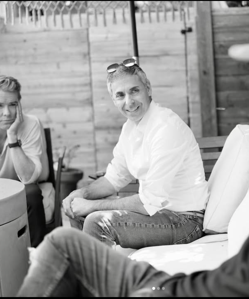 A man with sunglasses on his head smiling and sitting outdoors with wooden fencing in the background, while two women sit nearby in a casual setting.