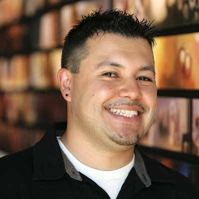 A smiling man with short, dark hair and a goatee standing in front of a blurred background of bookshelves.