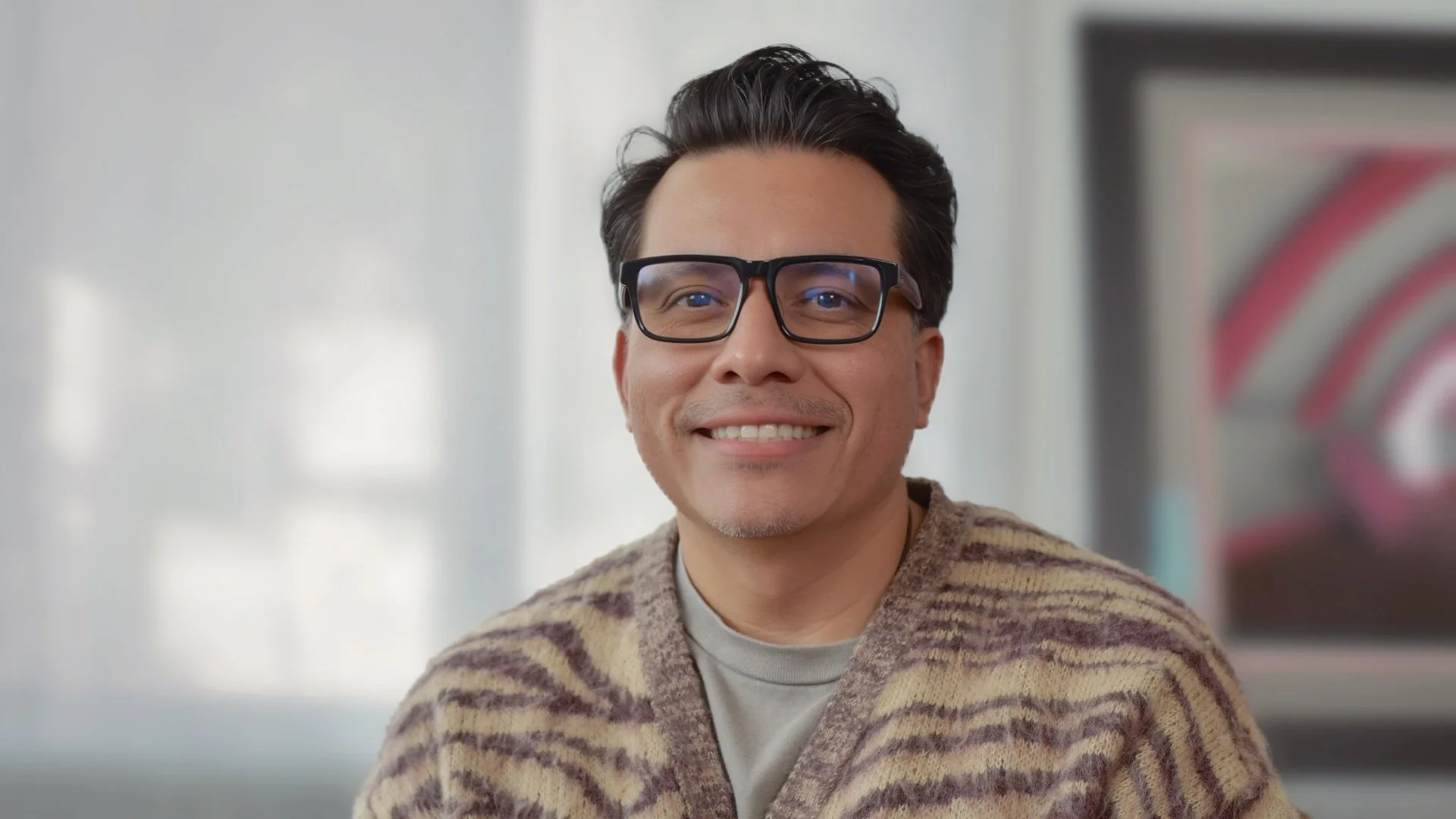 Smiling man with glasses and dark hair, wearing a patterned sweater, indoors with a blurred framed artwork in the background.