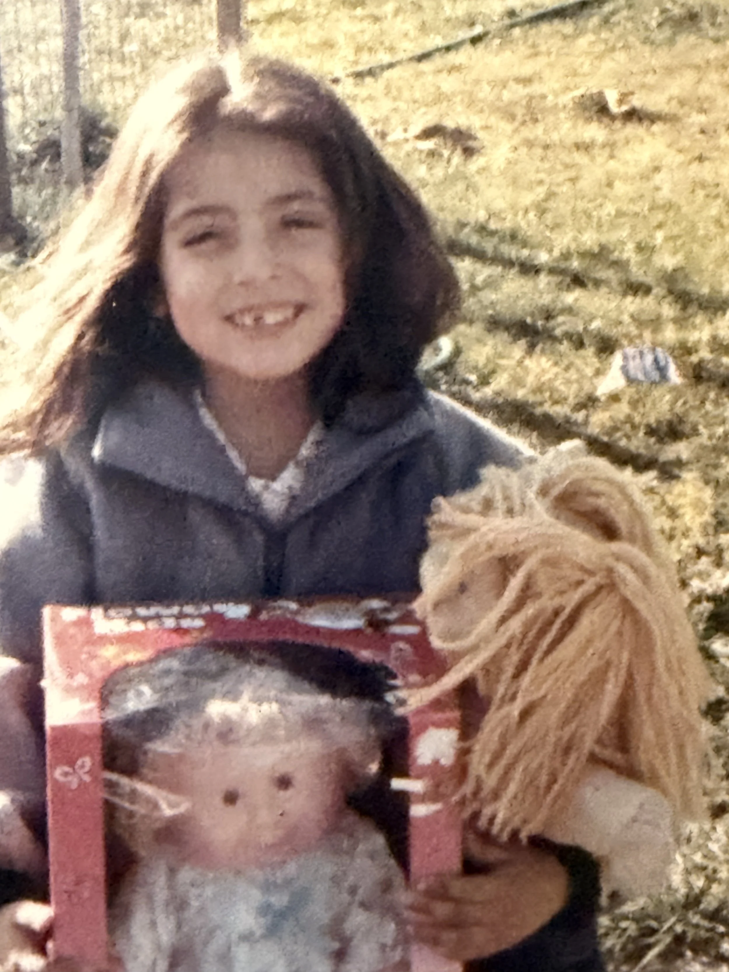 A smiling girl with brown hair in a ponytail, wearing a gray jacket, holding a dog with blonde yarn-like fur and a gift box with a teddy bear image on it, outdoors in a grassy area during daytime.