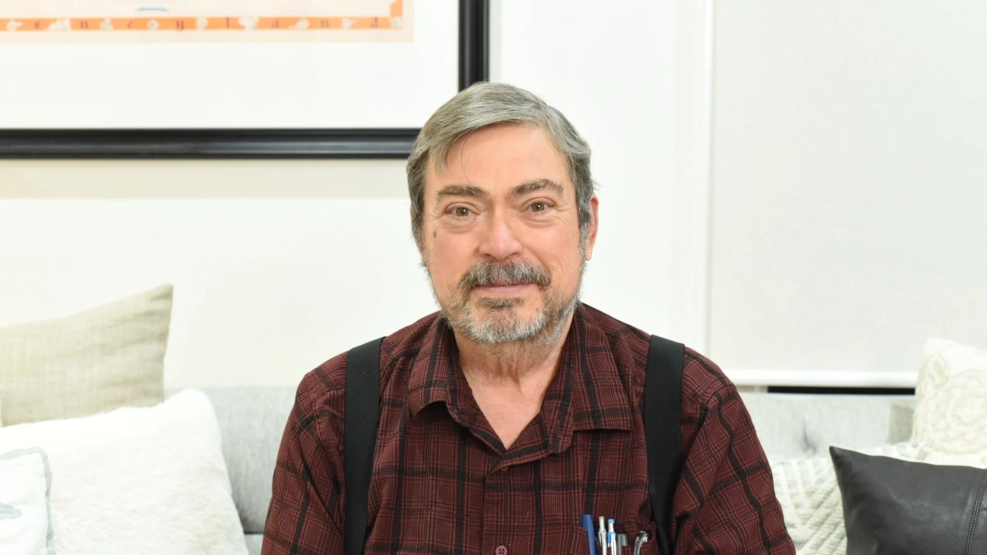 A middle-aged man with gray hair, beard, and wearing a plaid shirt with suspenders sitting in a room with white walls and pillows.