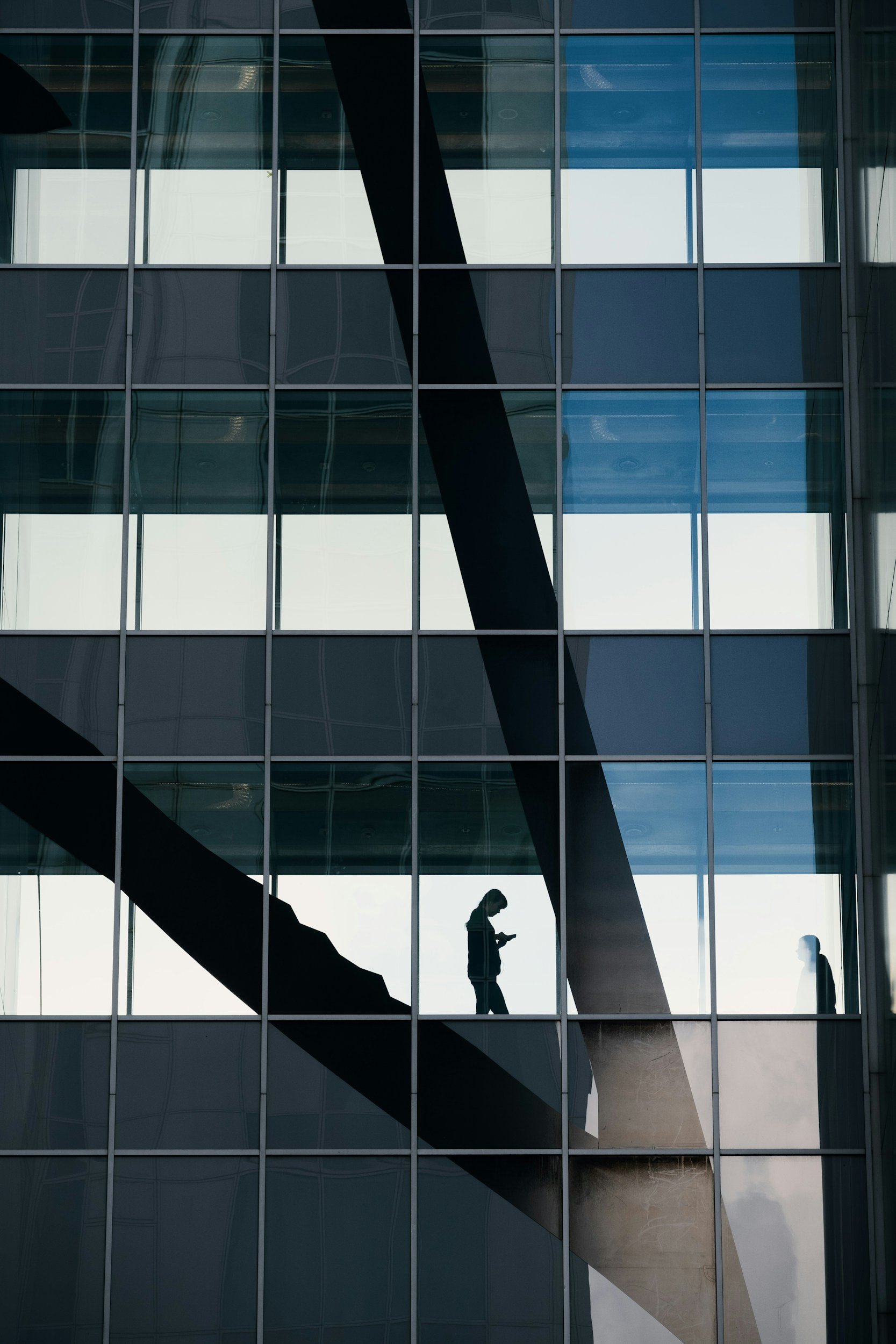 Silhouettes of two people on a glass building's interior walking and using a phone, with visible structural beams and window reflections.