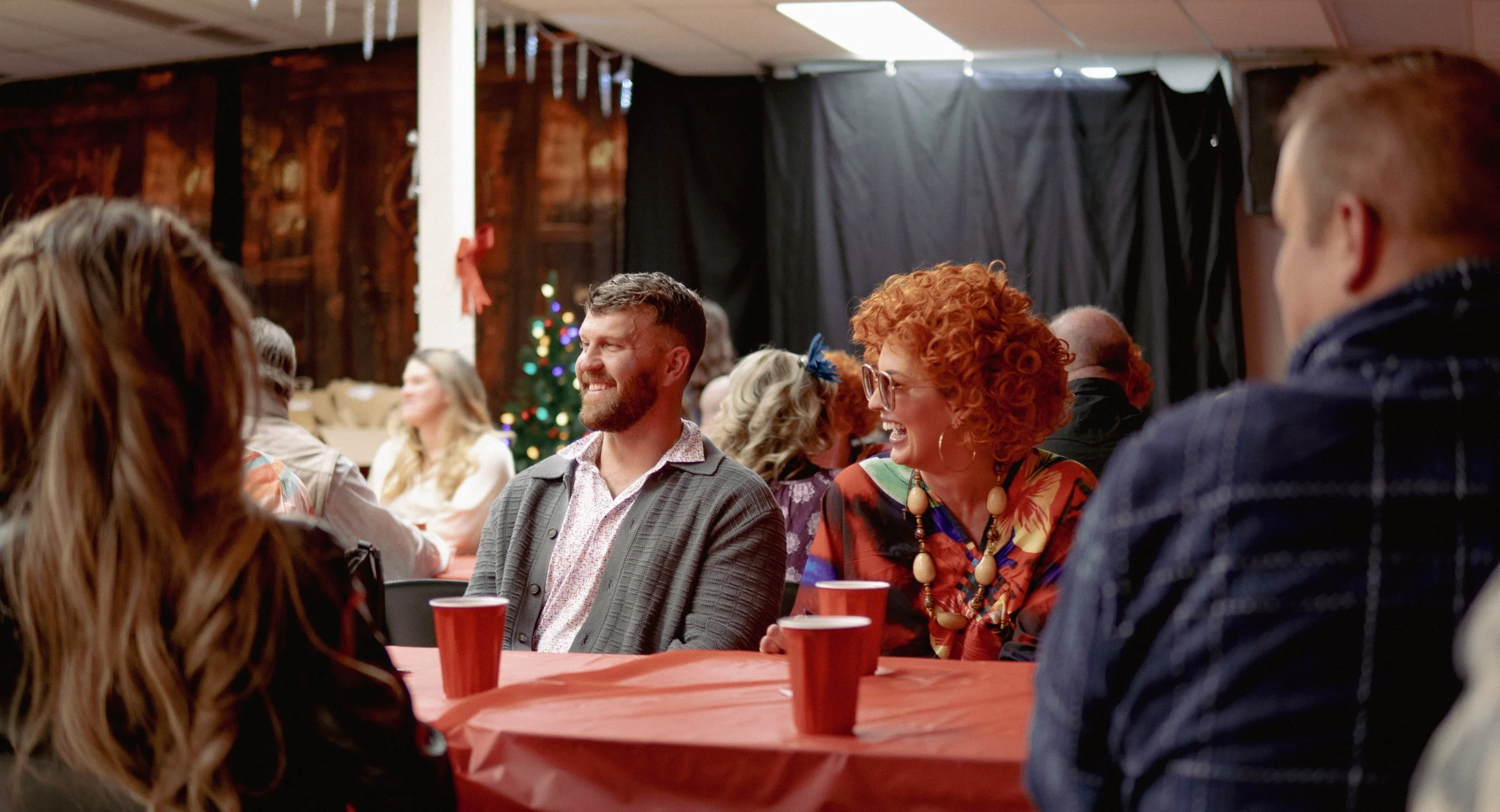 People sitting around a table at a holiday party, smiling and chatting, with a Christmas tree in the background.