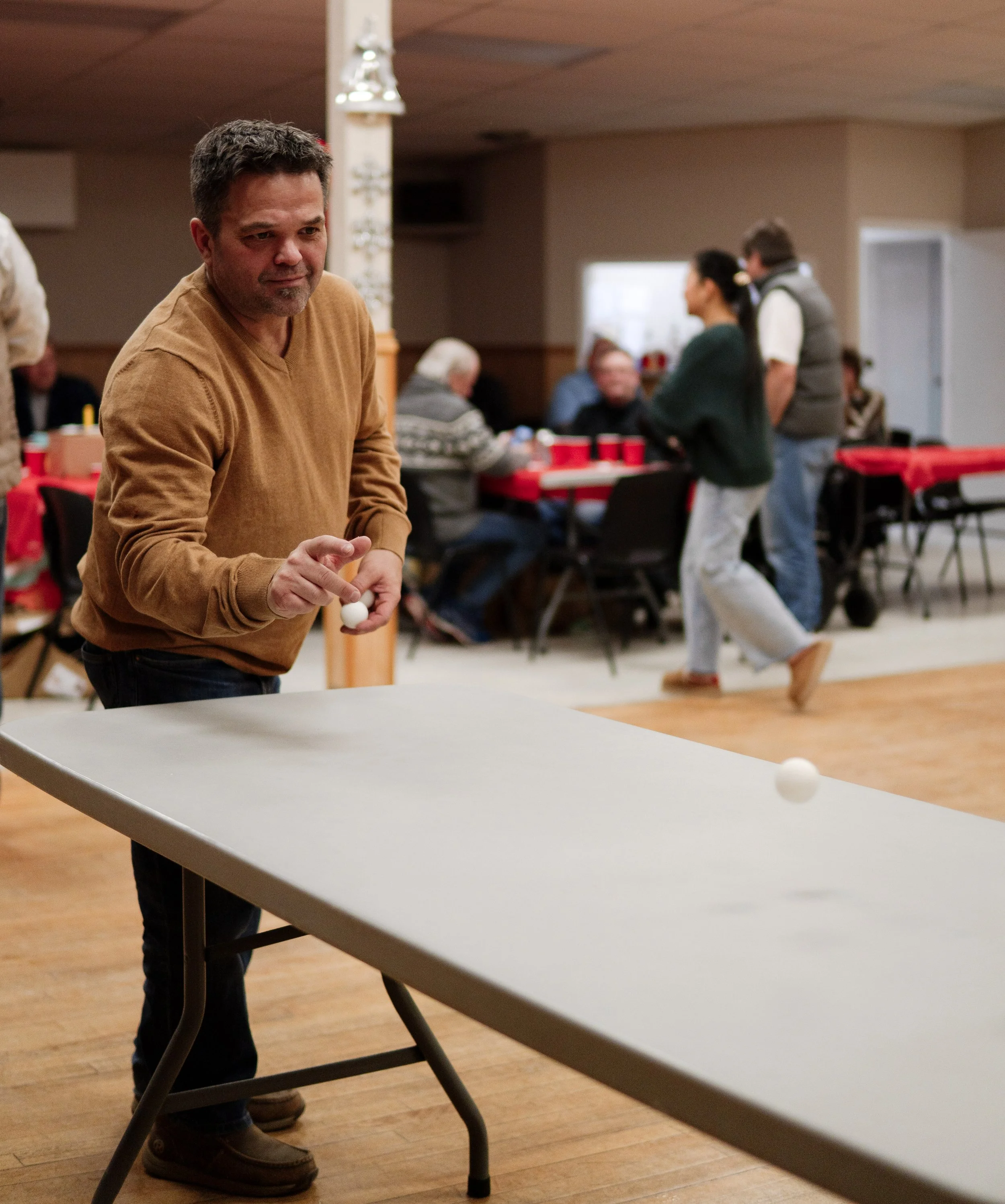 A man in a brown sweater playing ping pong at a social gathering in a community hall, with other people seated at tables and some standing in the background.