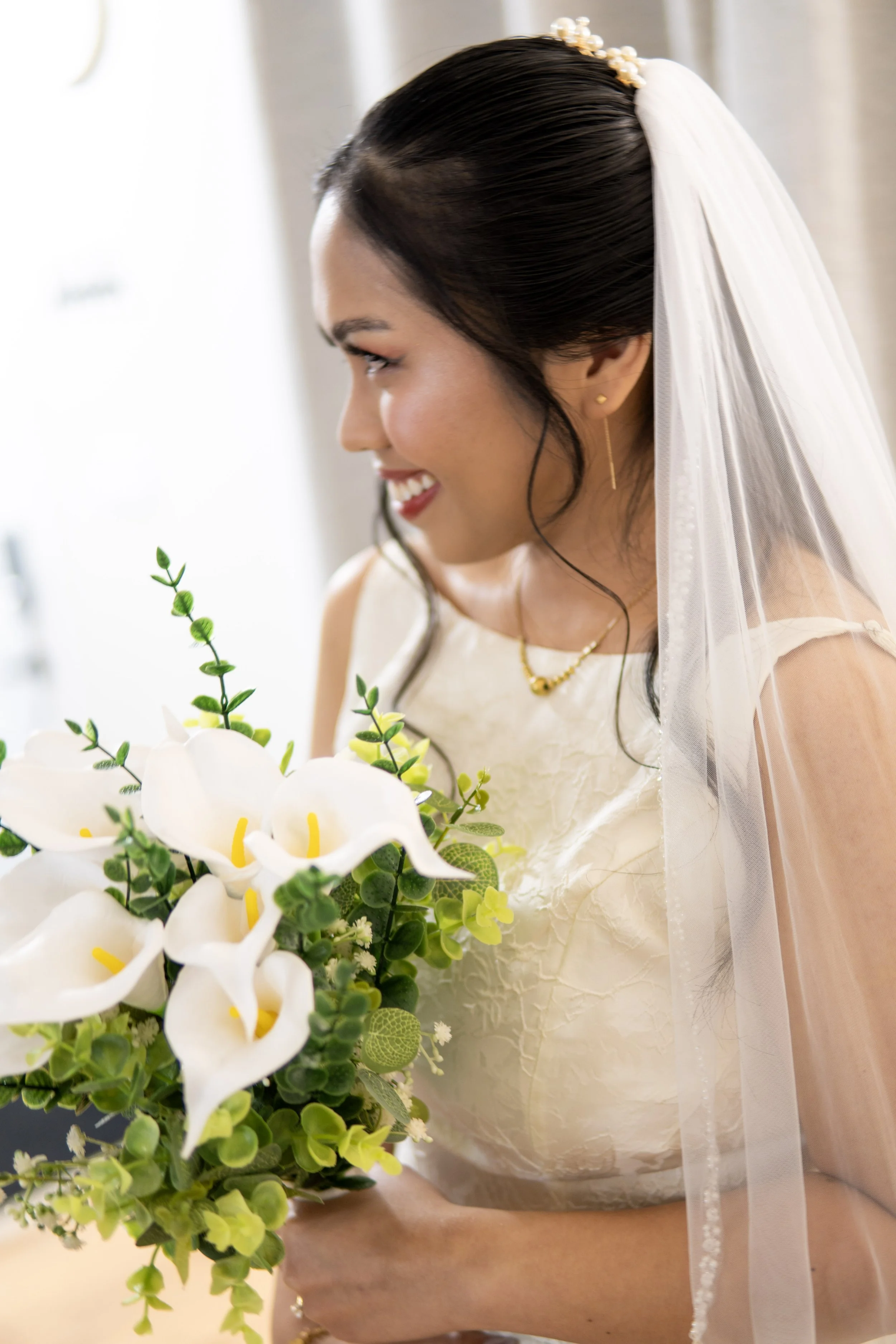 A bride with dark hair in a veil, holding a bouquet of white calla lilies and green foliage, smiling and looking to her right.