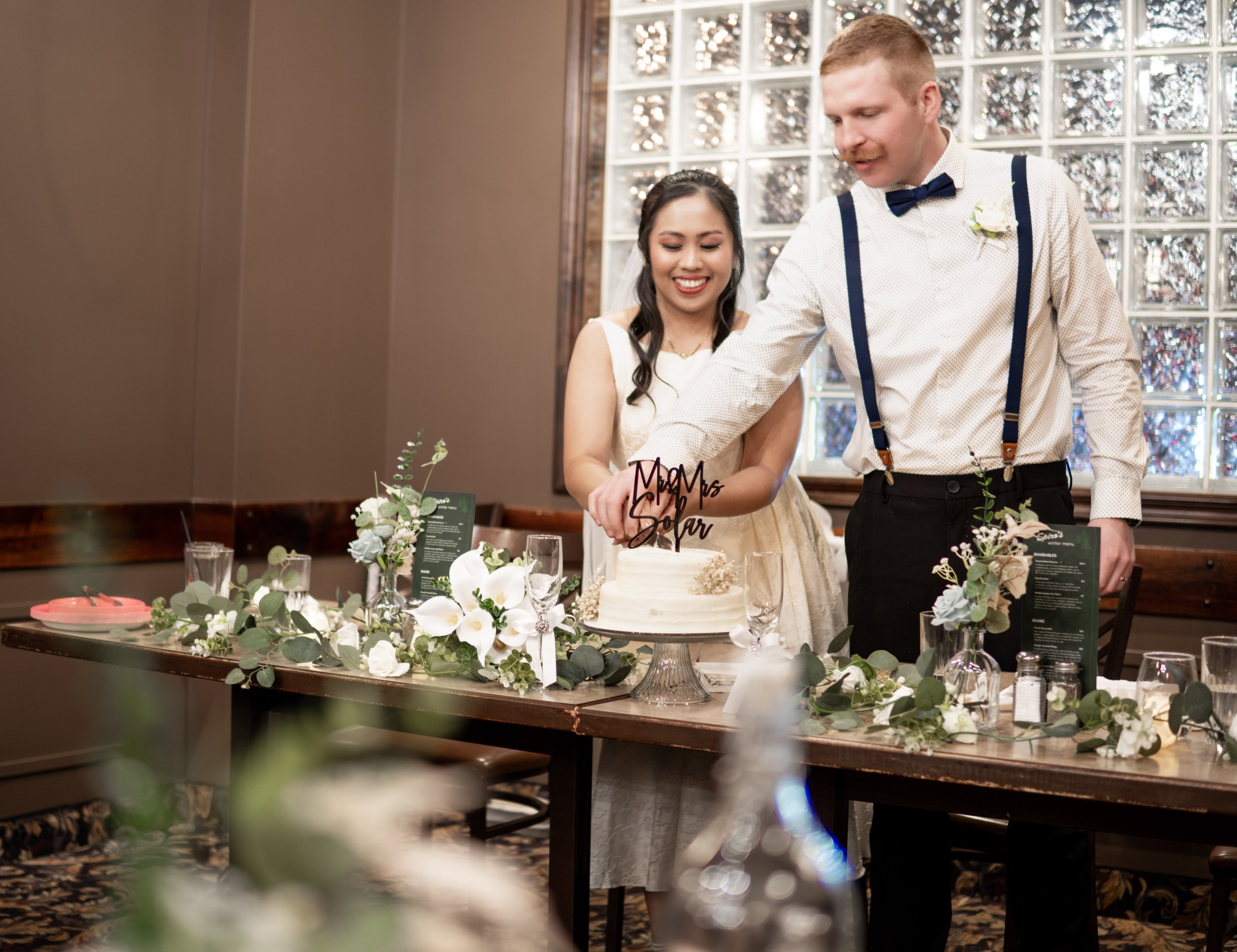 A bride and groom cutting a wedding cake together at their reception. The bride is smiling and wearing a white dress, while the groom is wearing a white shirt with navy suspenders and a bow tie. The table is decorated with flowers and greenery.