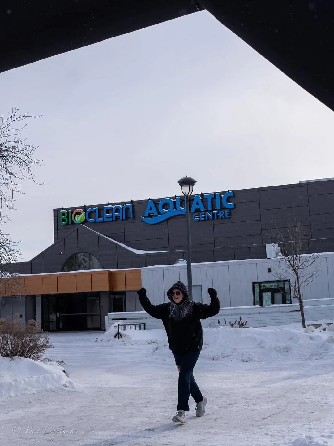 Person in black winter coat and sunglasses standing outdoors on snow, raising arms in front of a building with a sign that reads 'Bioclean Aquatic Centre.'