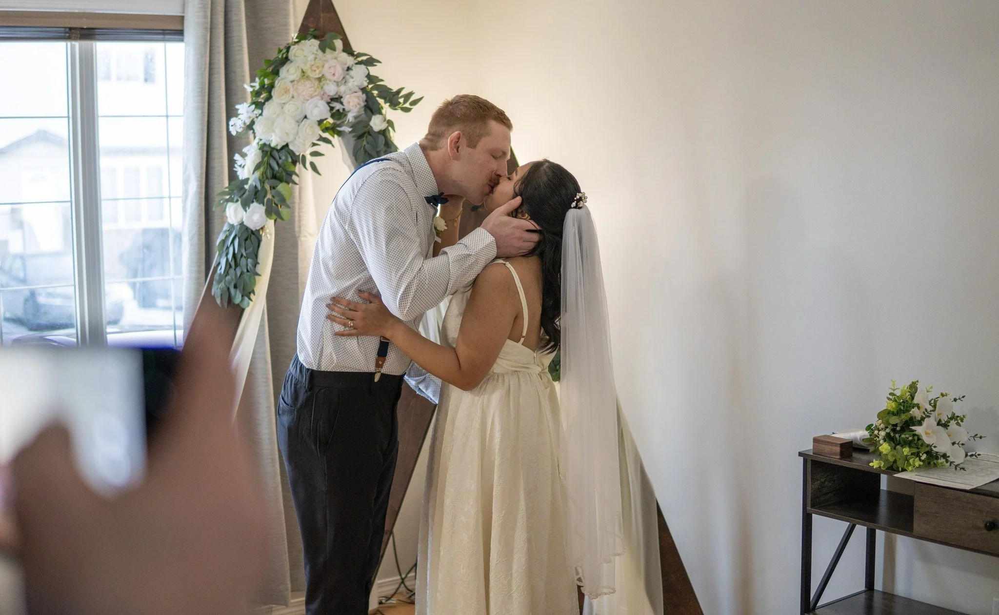 A couple sharing a kiss during a wedding ceremony in a bright room with a floral arch and minimal decor.