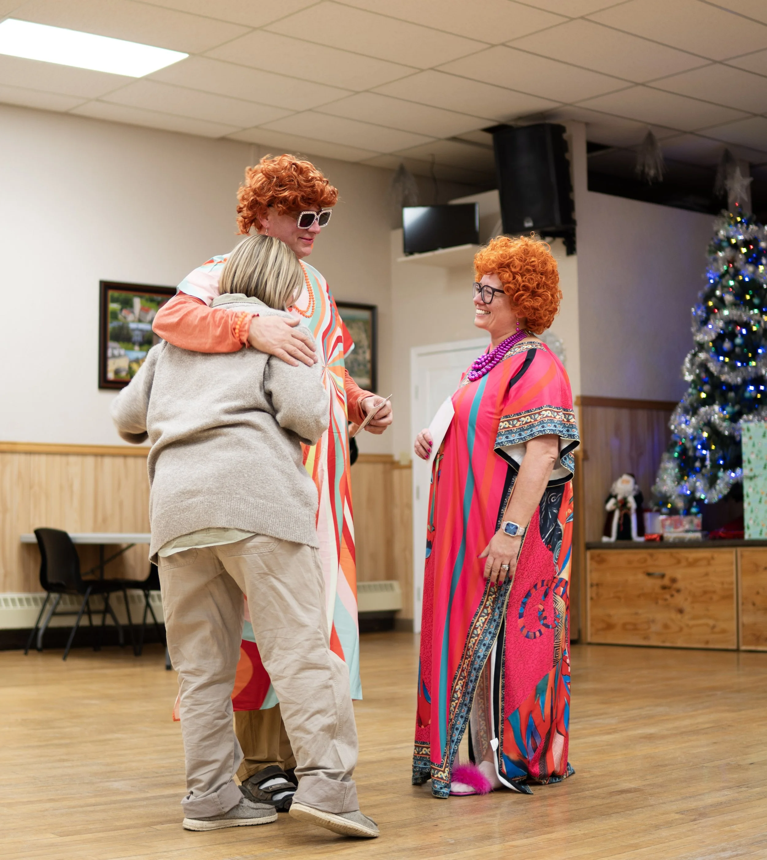 Three women, two dressed in colorful costumes with red curly wigs, are sharing a hug and smiling at each other in a room decorated for Christmas, with a decorated Christmas tree in the background.