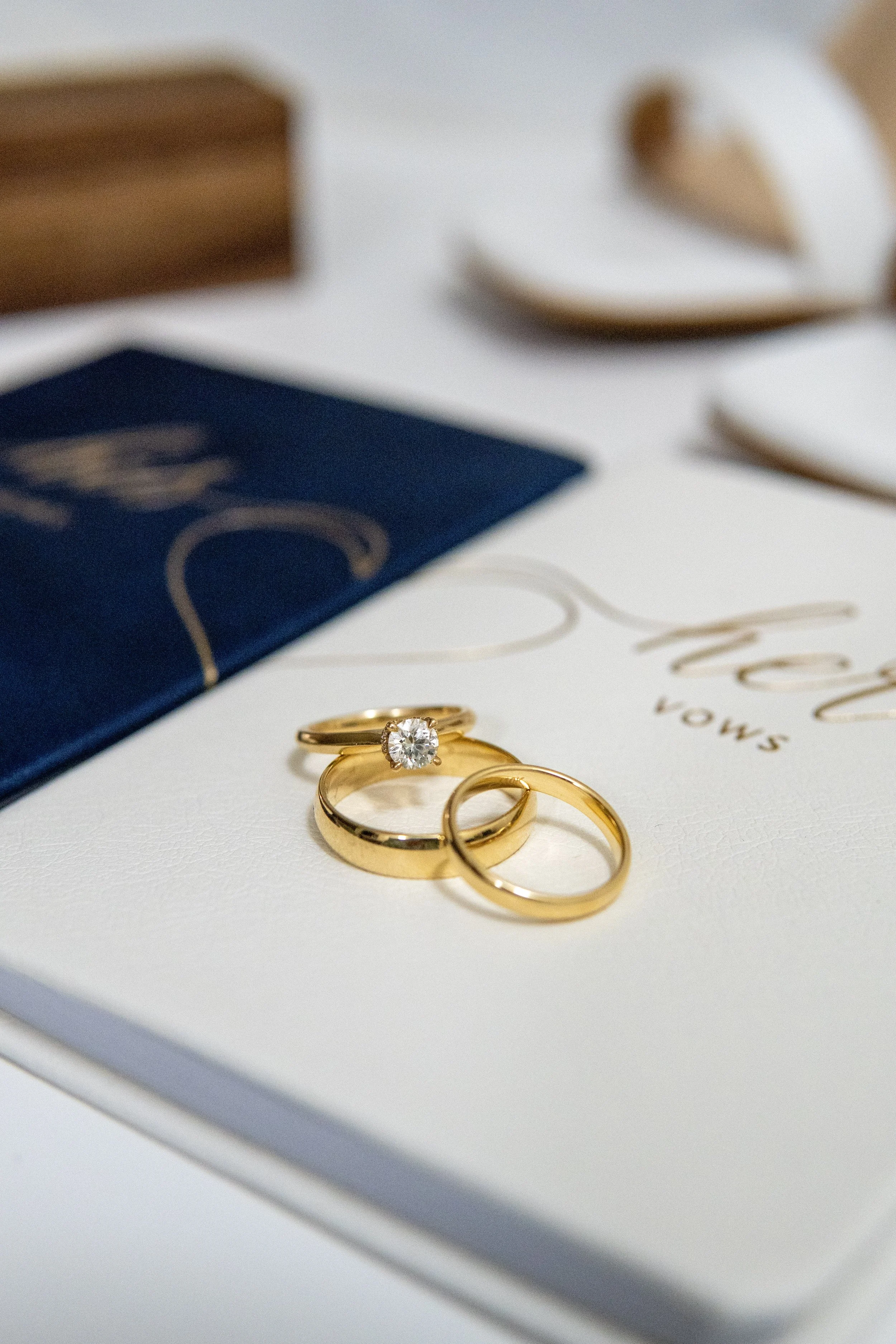Three gold rings, one with a diamond, resting on a white card with gold writing, surrounded by a navy box, wooden box, and white decorative objects.