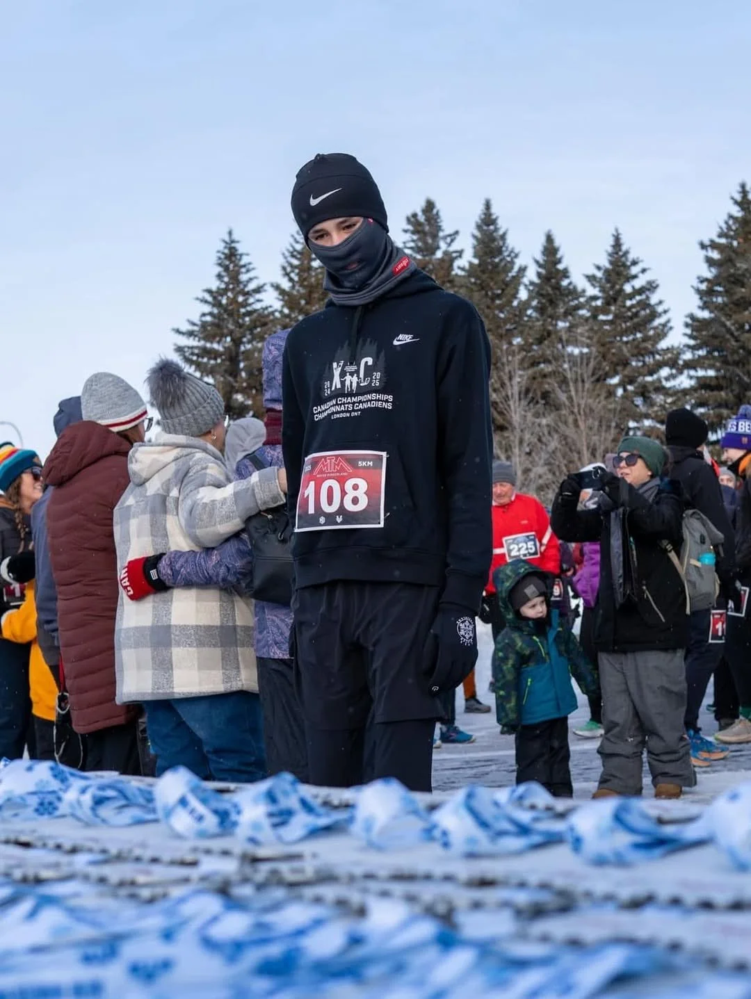 A young man dressed in black winter athletic gear, including a face covering and a beanie, stands among a crowd of people dressed for cold weather at an outdoor winter event, with some wearing race bibs, and snow-covered ground with blue and white clothing or banners in the foreground.