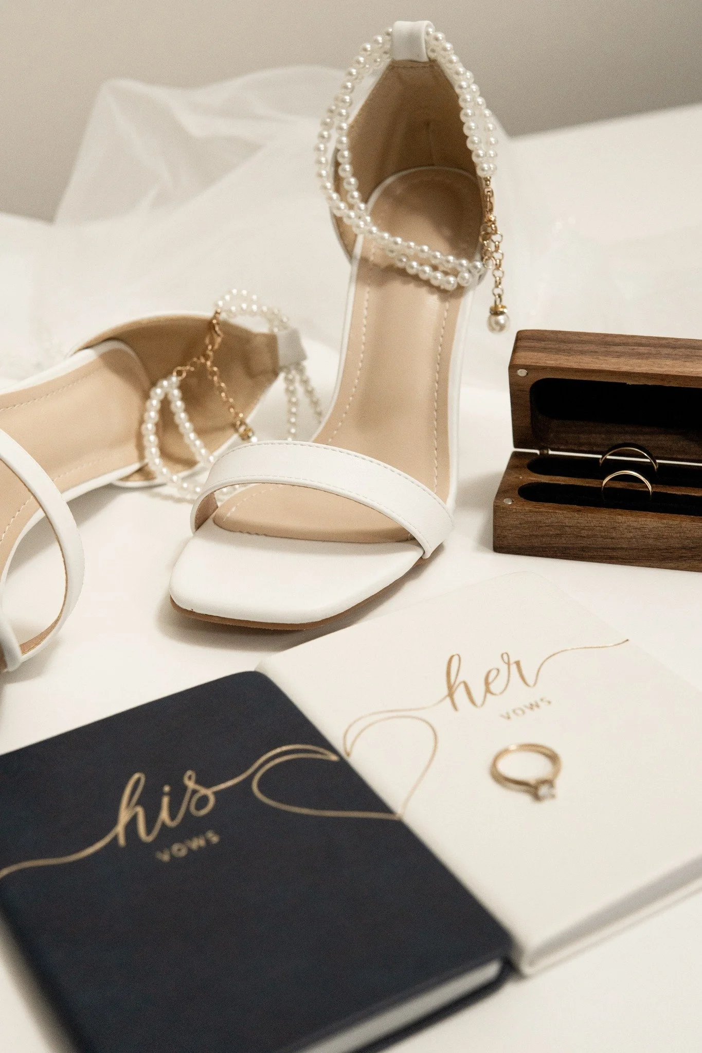 A pair of white high-heeled shoes decorated with pearl ankle straps. There are two vow books labeled 'his' and 'her' on a white surface, with a wedding ring placed on the 'her' vow book. A small wooden box with two rings inside is also visible.