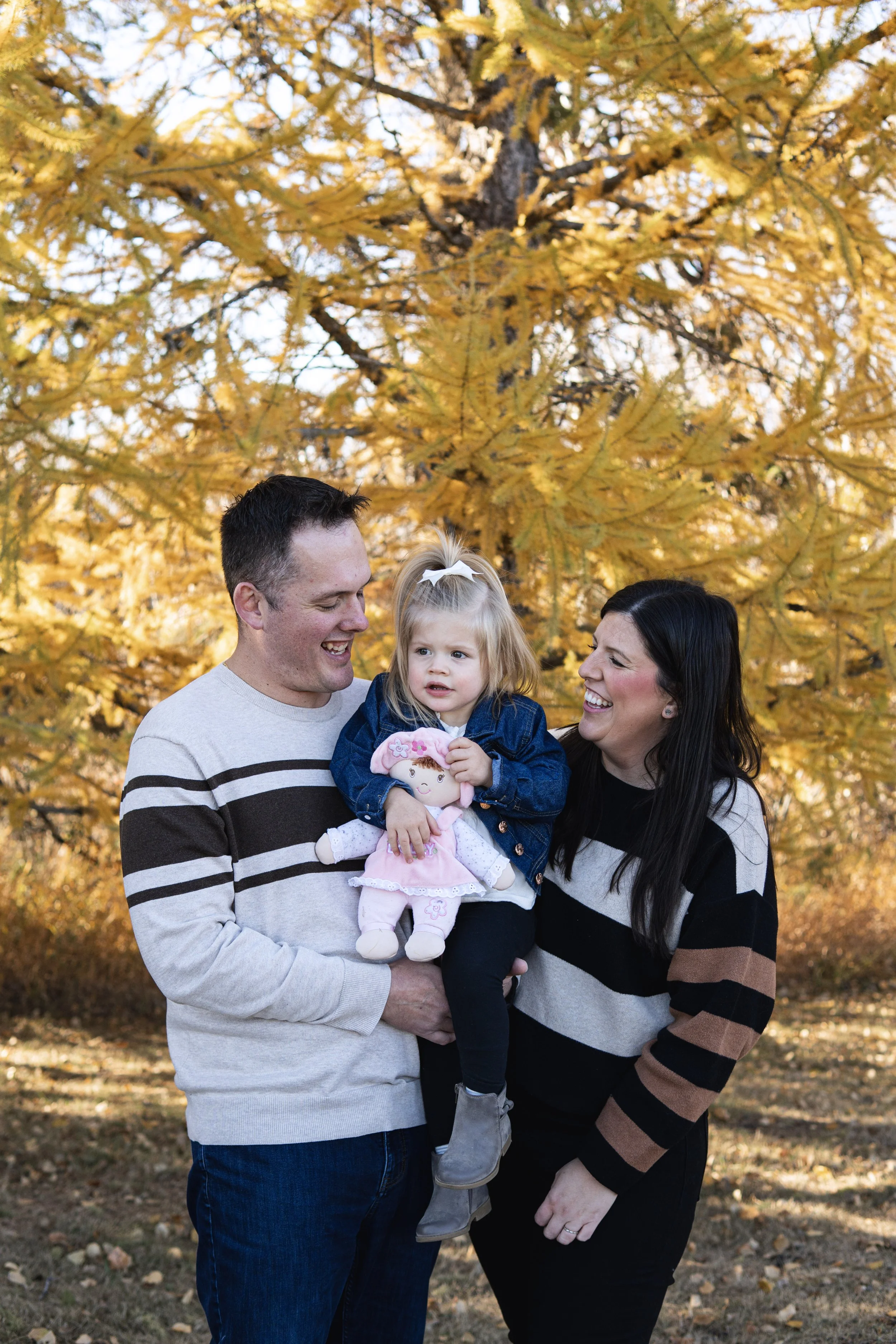 A family of three, a man, a woman, and a little girl, standing outside in fall with yellow-orange leaves on trees in the background. The man and woman are smiling at each other, while the girl is holding a doll and looking slightly confused.